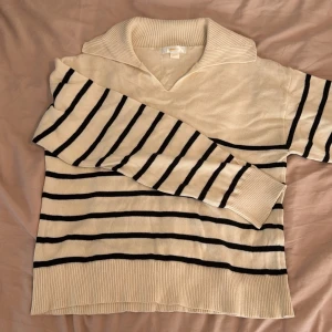 Randig krämvit tröja från Gina Tricot - Säljer en krämvit tröja från Gina Tricot med svarta horisontella ränder. Tröjan har en bred krage med v-ringning och ribbade muddar vid ärmslut och nederkant. Perfekt för en stilren och avslappnad look.