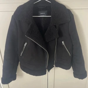 Svart teddyjacka från Stradivarius - Säljer en svart teddyjacka från Stradivarius med stora slag, dragkedjor och mysigt teddyfoder. Jackan har två snedställda fickor med dragkedja och en cool bikerstil. Perfekt för kyliga dagar när du vill vara både varm och snygg.