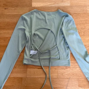 Topp med öppen rygg - Ljusgrön långärmad topp med ribbad struktur. Toppen har en öppen rygg med knytband och är croppad i modellen. Köpt second hand.