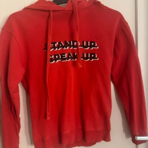 Röd hoodie med tryck 'Stand Up Speak Up' - Röd hoodie med huva och dragsnören. Tryck på bröstet med texten 'STAND UP SPEAK UP' i svart och vitt. Perfekt för en avslappnad stil och enkel att matcha med jeans eller joggers.
