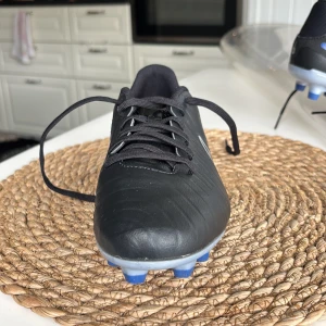 Nike Tiempo svart fotbollsskor  - Säljer en svart Nike Tiempo fotbollssko med blå och vita detaljer. Skon har snörning och är tillverkad i syntetmaterial med en platt sula och dobbar för spel på gräs. Klassisk Nike-logga på sidan och blå inslag vid hälen.