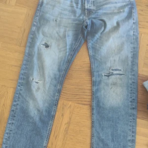 Blå slitna jeans från Jack & Jones - Säljer ett par blå jeans från Jack & Jones med slitna detaljer på benen. Jeansen har klassisk femficksdesign, normal passform och är tillverkade i denim. Perfekta för en avslappnad stil.