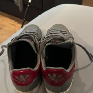 Grå Adidas Stan Smith sneakers med röda detaljer - Säljer ett par grå Adidas Stan Smith sneakers med röda hälpaneler och vita sulor. Klassisk låg modell med snörning och diskreta perforerade ränder på sidorna. Perfekta för dig som gillar stilrena och sportiga skor.