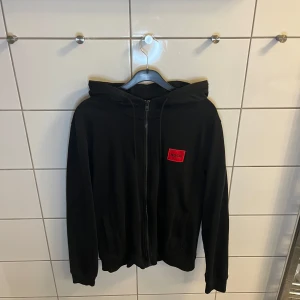 Hoodie - Hugo Boss - Clean hoodie från Hugo Boss med dragkedja framtill och röd logotyp på bröstet. Tröjan har huva och långa ärmar, perfekt för en avslappnad stil. Klassisk och enkel design som passar till mycket.