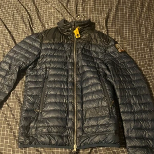 Mörkblå dunjacka från Parajumpers - Säljer en mörkblå dunjacka från Parajumpers med dragkedja och hög krage. Jackan har quiltad design, två sidofickor med dragkedja och en patch på ärmen. Insidan är ljusgrå med orange detaljer. Perfekt för sommaren