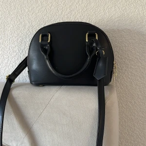 Svart handväska med axelrem - Snygg svart handväska med både handtag och justerbar axelrem. Väskan har guldfärgade metalldetaljer och dragkedja upptill. Insidan är fodrad i vinrött tyg och har små innerfack för organisering. Perfekt storlek för det viktigaste. Allt är i bra skick köpte ifrån Shein. Säljer den då den inte kommer nu för tiden till användning. Vid fler bilder hör av er ☺️
