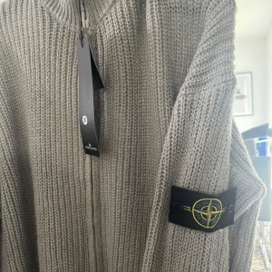 Grå stickad kofta från Stone Island - Säljer en grå stickad kofta från Stone Island med dragkedja framtill och klassisk logotyp på ärmen. Koftan har lång ärm och är ribbstickad för en snygg och modern look. Perfekt för lager-på-lager-stil. Passar L/Xl