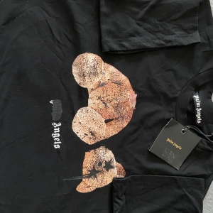 Svart t-shirt med nalletryck från Palm Angels - Svart t-shirt från Palm Angels med stort tryck av en nalle på framsidan. Klassisk passform med rund hals och korta ärmar. Den är lite för stor för mig som tar storlek S så det passar M och S bra.
