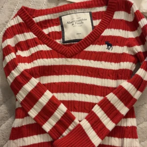 Rödvit randig ribbad tröja från Abercrombie & Fitch - Säljer en ribbad långärmad tröja från Abercrombie & Fitch i rödvitt randigt mönster. Tröjan har v-ringning och en liten blå broderad älg på bröstet. Perfekt för dig som gillar klassisk och färgglad stil.