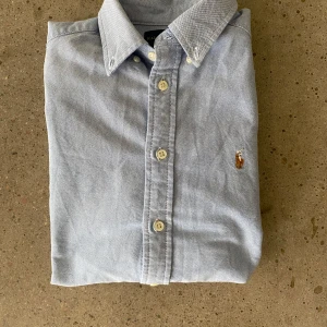 Ljusblå skjorta från Ralph Lauren - Klassisk ljusblå skjorta från Ralph Lauren med button-down krage och broderad logga på bröstet. Skjortan har långa ärmar och knäppning framtill. Perfekt för en stilren och tidlös look. Storlek xs