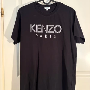 Svart t-shirt från Kenzo - Säljer en svart t-shirt från Kenzo med stort vitt tryck 'KENZO PARIS' på bröstet. Klassisk rund halsringning och korta ärmar. Perfekt för dig som gillar stilrena och trendiga plagg med tydlig logga.
