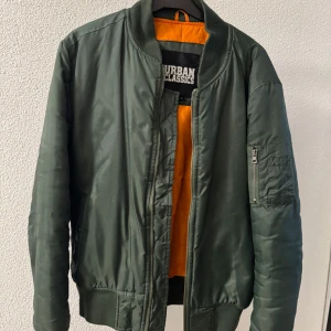 Grön bomberjacka från Urban Classics - Säljer en klassisk grön bomberjacka från Urban Classics med orange foder. Jackan har dragkedja framtill, ribbade muddar och en ficka med dragkedja på ärmen. Perfekt för dig som gillar streetwear.