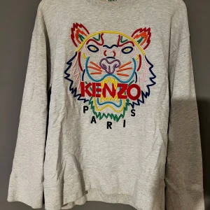 KENZO Grå sweatshirt med tigerbrodyr  - Säljer en grå sweatshirt från Kenzo med ikonisk tigerbrodyr och färgglada detaljer framtill. Tröjan har rund halsringning och långa ärmar. Perfekt statement-plagg för dig som gillar streetwear och vill sticka ut.