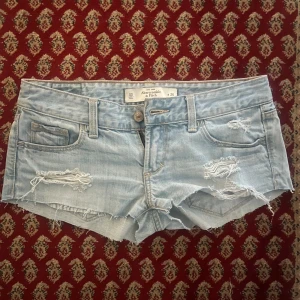 Ljusblå jeansshorts från Abercrombie & Fitch - Säljer ett par ljusblå jeansshorts från Abercrombie & Fitch i storlek W24. Shortsen har slitna detaljer, fransig kant och klassisk femficksdesign. Perfekta för sommaren och har låg midja för en avslappnad look.