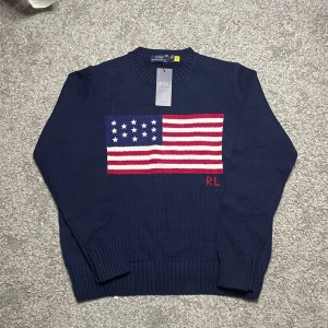 Ralph Lauren tröja med flaggtryck (Iconic Flag Sweater)  - Ralph Lauren tröja med flaggtryck (Iconic Flag Sweater) 🇺🇸 – M  Säljer en riktigt snygg och klassisk Ralph Lauren-tröja med det ikoniska amerikanska flaggtrycket framtill! Köpte den begagnad till min brorsa, men han ville inte ha den. Den tidigare ägaren hade aldrig använt den – alltså i princip oanvänd. ✅ QR-koden fungerar som den ska, vilket ofta tyder på äkthet.  • Storlek: M • Skick: nyskick med tagen kvar • Design: Flaggtryck (Iconic Flag Sweater) • Märke: Polo Ralph Lauren • Material: Mjukt