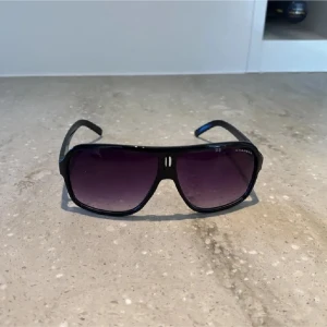 Svarta solglasögon från Carrera - Snygga svarta solglasögon från Carrera med stora, rektangulära bågar och mörka tonade glas. Bågarna är i plast och har Carrera-loggan på sidan. Perfekt accessoar för en cool look. Pris kan diskuteras vid snabb affär.