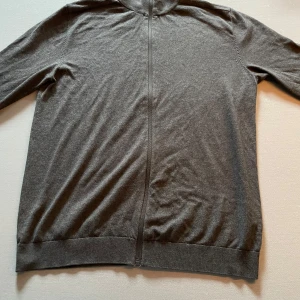 Grå ziptröja i bomull - En stilren grå ziptröja med hel dragkedja och ståkrage. Tröjan är tillverkad i mjuk bomull och har långa ärmar samt ribbade muddar vid ärmslut och nederkant. Perfekt för lager-på-lager och enkel att matcha med andra plagg. 