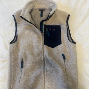 Beige fleeceväst från Patagonia - Mysig beige fleeceväst från Patagonia med marinblå detaljer och dragkedja framtill. Västen har en hög krage och en praktisk bröstficka med dragkedja. Perfekt lager.