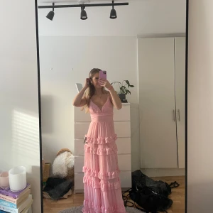 Ljusrosa långklänning med volanger - Säljer en ljusrosa långklänning med smala axelband och djup v-ringning. Klänningen har flera lager volanger och markerad midja. Perfekt för bal! Klänningen är helt oanvänd och prislappen är kvar. Jag säljer på grund av att jag har hittat en annan klänning till balen. Köpt för 1300kr
