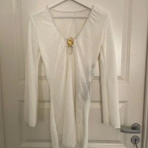 Vit strandklänning med gulddetalj - Säljer en vit långärmad klänning med v-ringning och  guldfärgad snäcka som detalj framtill. Klänningen är i ett lätt, halvtransparent syntetmaterial. 