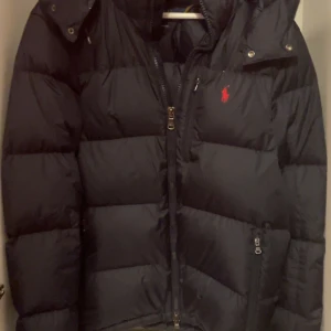  Polo Ralph Lauren down jacket blå - Säljer en svart dunjacka från Polo Ralph Lauren med huva och dragkedja. Jackan har en klassisk pufferdesign och röd broderad logga på bröstet. Perfekt för kyliga dagar och riktigt snygg till vardags.