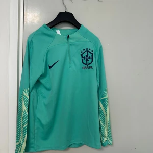 Brasilien långärmad fotbollströja - Snygg långärmad Brasilien fotbollströja i turkos med mörkblå Nike-logga och Brasilien-emblem på bröstet. Ärmarna har ljusgula grafiska detaljer. Perfekt för dig som gillar fotboll och vill sticka ut på planen.