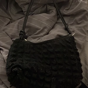 Svart axelväska med struktur - Säljer en svart axelväska med mjuk, strukturerad yta. Väskan har dragkedja upptill och guldfärgade metalldetaljer. Insidan är fodrad i beige tyg. Perfekt för dig som vill ha en stilren men unik accessoar. Får plats med relativt mycket saker