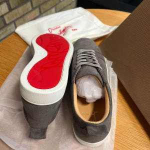 Gråa sneakers från Christian Louboutin - Säljer ett par gråa sneakers från Christian Louboutin med klassisk röd sula. Skorna har snörning, rund tå och är tillverkade i mocka med vita gummisulor. Perfekta för dig som gillar stilrena och exklusiva sneakers. Storlek 42 men passar 41