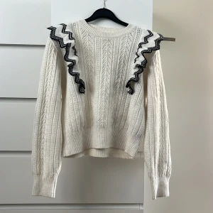 Vit stickad tröja Lindex - Säljer en vit stickad tröja från Lindex med snygga volangdetaljer längs axlarna, dekorerade med svart sicksackmönster. Tröjan har lång ärm och rund halsringning. Perfekt för dig som gillar unika detaljer!
