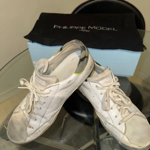 Vita sneakers från Philippe Model - Säljer ett par vita Philippe Model sneakers med grå detaljer och svarta inslag vid hälen. Skorna har snörning och en platt sula. Klassisk design med rund tå och diskret logotyp på sidan. Perfekta för en stilren vardagslook.