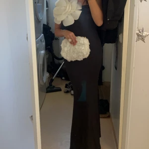 Svart långklänning med stora vita blommor - Säljer en svart långklänning med figurnära passform och stora vita blommor som dekoration vid axel och midja. Klänningen har ärmlös design och en elegant siluett som verkligen sticker ut.