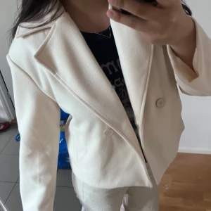 Beige kappa - Säljer en stilren beige kappa med två knappar framtill och klassisk krage. Jackan har lång ärm och är tillverkad i ett mjukt material som ger en snygg och clean look. Perfekt för dig som vill ha en enkel men trendig kappa. Perfekt till våren och sommaren.