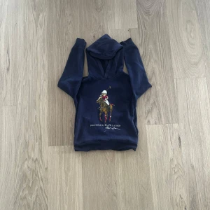 Mörkblå hoodie från Polo Ralph Lauren - Säljer en mörkblå hoodie från Polo Ralph Lauren med stort tryck av en polospelare på häst framtill. Klassisk huva och mjukt material, perfekt för en avslappnad stil. Ikonisk logga och snygga detaljer.