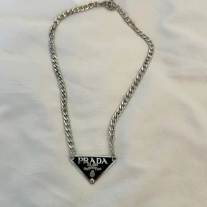 Silverfärgat halsband från Prada - Säljer ett snyggt halsband från Prada i silverfärg med en kraftig kedja och en ikonisk svart triangelplatta med logotypen 'PRADA MILANO'. Perfekt statement-piece för att lyfta din outfit.