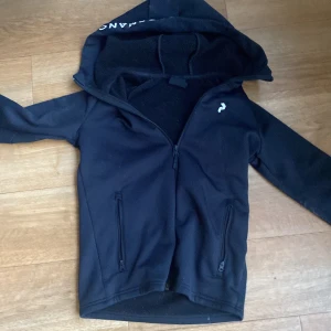Svart hoodie från peak performance - Svart hoodiejacka från Röhnisch med dragkedja framtill och två fickor med dragkedja. Jackan har en stor huva och en liten vit logga på bröstet. Perfekt för träning eller som ett extra lager.