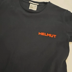 Svart t-shirt från Helmut Lang med röd text - Svart t-shirt från Helmut Lang med röd text 'HELMUT' på bröstet och stort tryck 'IN LANG WE TRUST' på ryggen. Tillverkad i 100% bomull och har en klassisk passform med korta ärmar. Perfekt för dig som gillar stilrena statement-plagg.