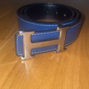  H bälte  - Säljer ett stilrent blått bälte med ett stort H-format metallspänne. Bältet har vita sömmar längs kanterna och är gjort i skinn. Perfekt accessoar för att lyfta din outfit.