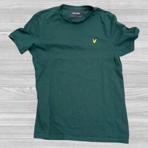 Lyle & Scott t-shirt - Mörkgrön t-shirt från Lyle & Scott med klassisk passform och korta ärmar. T-shirten har en gul broderad logga på bröstet och är tillverkad i 100% ekologisk bomull. Perfekt för en stilren och enkel look.