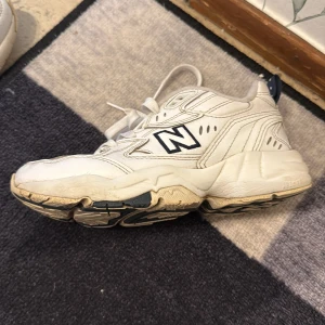 Vita New Balance 608 sneakers - Säljer ett par klassiska vita New Balance 608 sneakers med blå detaljer och tjock sula. Skorna har snörning och är perfekta för en sportig och avslappnad stil. Ikonisk N-logga på sidan och bekväm passform.