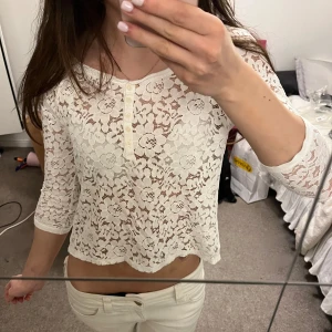 Vit spetsblus med trekvartsärm - Säljer en söt vit blus i spets med blommigt mönster och trekvartsärm. Blusen har rund halsringning, knappar framtill och är lätt croppad. Perfekt till sommaren!