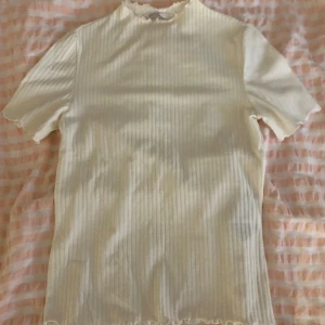 Vit ribbad topp från ONLY - Säljer en vit ribbad topp från ONLY med korta ärmar och liten volangkant vid hals och nederkant. Toppen har en tight passform och är perfekt för en stilren look.