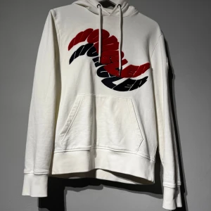 Moncler hoodie - Säljer en vit hoodie från Moncler med ett stort grafiskt tryck i rött och svart på bröstet. Tröjan har huva med dragsko och en stor magficka framtill. Perfekt för dig som gillar exklusiva märken. Skriv om du har några funderingar. Självklart äkta.