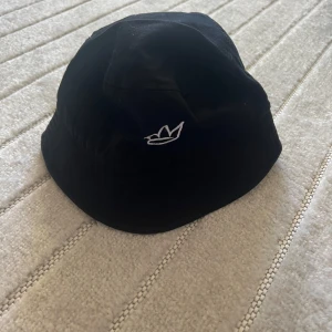Svart bucket hat med broderad krona - Snygg svart bucket hat med vit broderad krona framtill. Hatten har klassisk form med mjuk brätte och är tillverkad i ett lätt och bekvämt material. Perfekt accessoar för en stilren look.