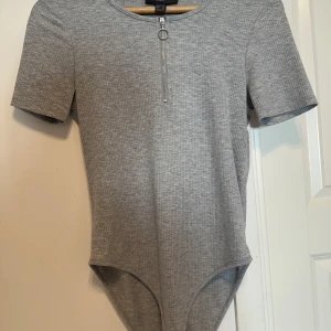 Grå ribbad bodysuit från Primark - Säljer en grå ribbad bodysuit från Primark med korta ärmar och rund halsringning. Framtill finns en dekorativ dragkedja med ringdetalj. Perfekt att styla med jeans eller kjol. Den är använd några gånger men är i ett jättebra skick. 