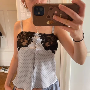 Vit och svart linnetopp med spets - Säljer en vit linnetopp med tunna svarta ränder och svarta spetsdetaljer upptill. Toppen har smala axelband och knappar framtill. Perfekt för sommaren och ger en elegant touch till din outfit.