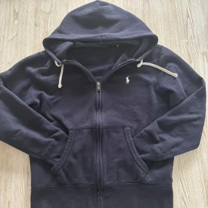 Svart hoodie från Polo Ralph Lauren - Klassisk svart hoodie från Polo Ralph Lauren med dragkedja framtill och vit broderad logga på bröstet. Tröjan har huva med vita snören och två fickor framtill. Perfekt för en avslappnad stil. Storlek S 