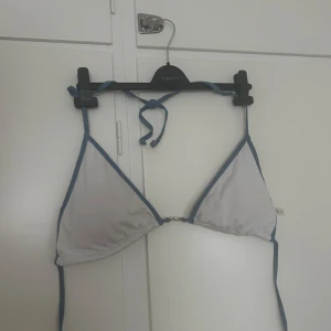 Vit trekantsbikini med blå kant - Säljer en vit trekantsbikini med blå kant och knyt i nacken. Klassisk modell med tunna band och enkel design, perfekt för stranden eller poolen.