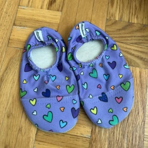 Lila tofflor med hjärtmönster - Säljer ett par lila tofflor med färgglada hjärtan i olika nyanser. De har mjuk, ljusgrå sula och resår runt öppningen för enkel påtagning. Perfekta för inomhusbruk och supergulliga till barn.
