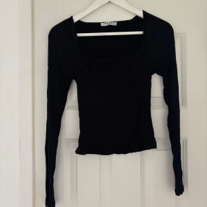Svart ribbad topp från NA-KD - Svart långärmad topp från NA-KD med fyrkantig urringning och ribbad struktur. Sitter snyggt och har en slim passform som framhäver figuren. Perfekt att styla med jeans eller kjol för en clean look.