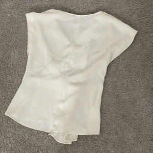 Vit omlottopp i satin från  Zara - Säljer en stilren vit omlottopp från zarai glansigt satinmaterial. Toppen har korta ärmar, en snygg twist framtill och en lätt v-ringning. Perfekt för dig som gillar en clean och chic look med lite extra detalj.